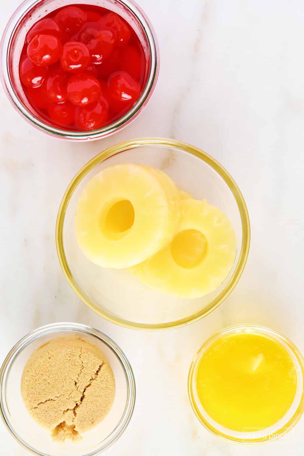 Pineapple upside down cake topping ingredients on a marble surface: pineapple slices, butter, brown sugar, and cherries.