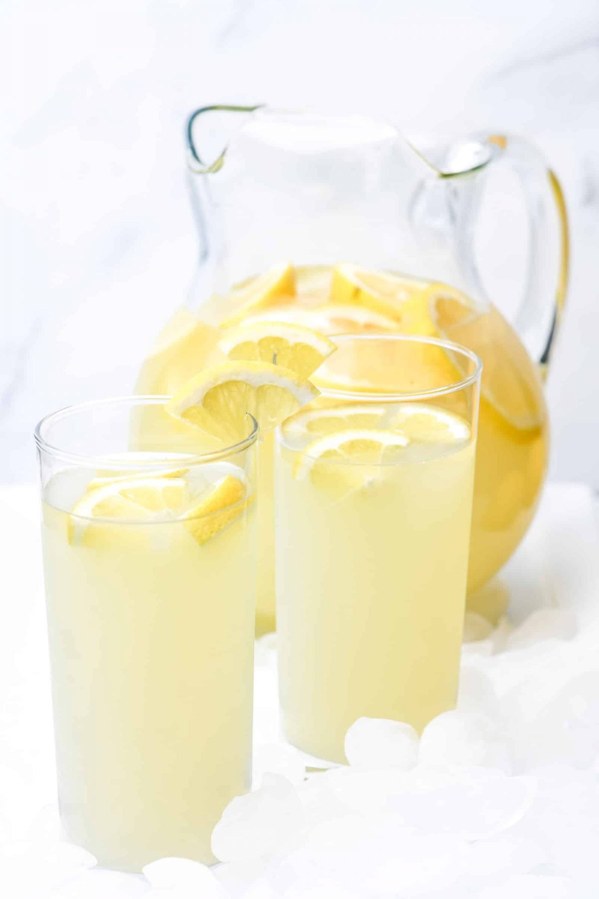 Pitcher of lemonade with fresh lemon slices flanked by two glasses with lemonade topped with lemon slices are all sitting on tray of ice cubes.