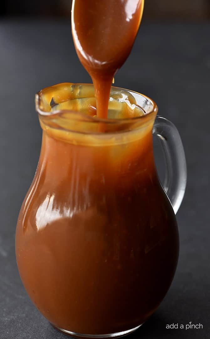 Salted Caramel Sauce in a glass pitcher and being drizzled into pitcher with a spoon. 