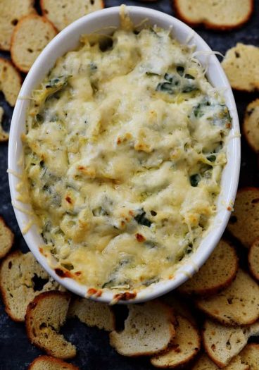 Hot Spinach Artichoke Dip in a white oval bowl surrounded by bagel chips // addapinch.com
