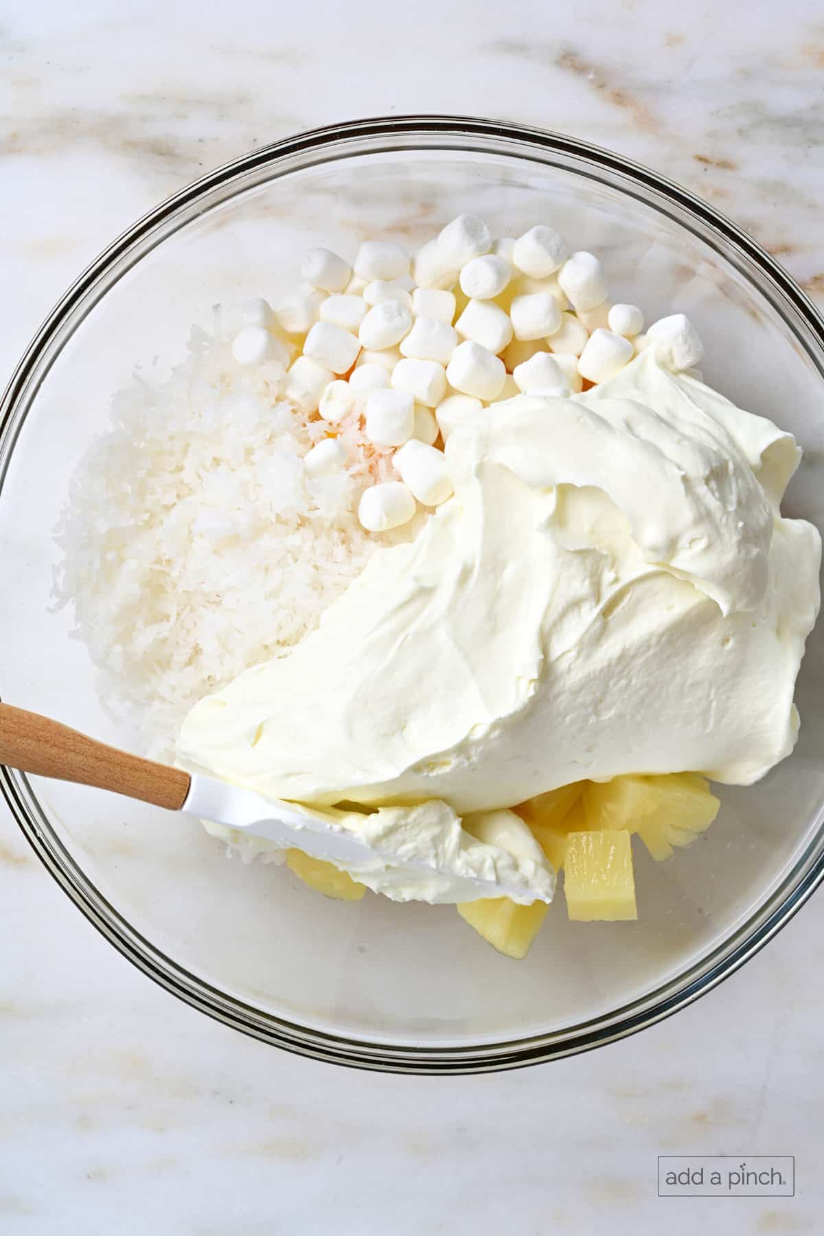 Fruit, marshmallows, coconut, and creamy dressing in a glass mixing bowl for ambrosia.