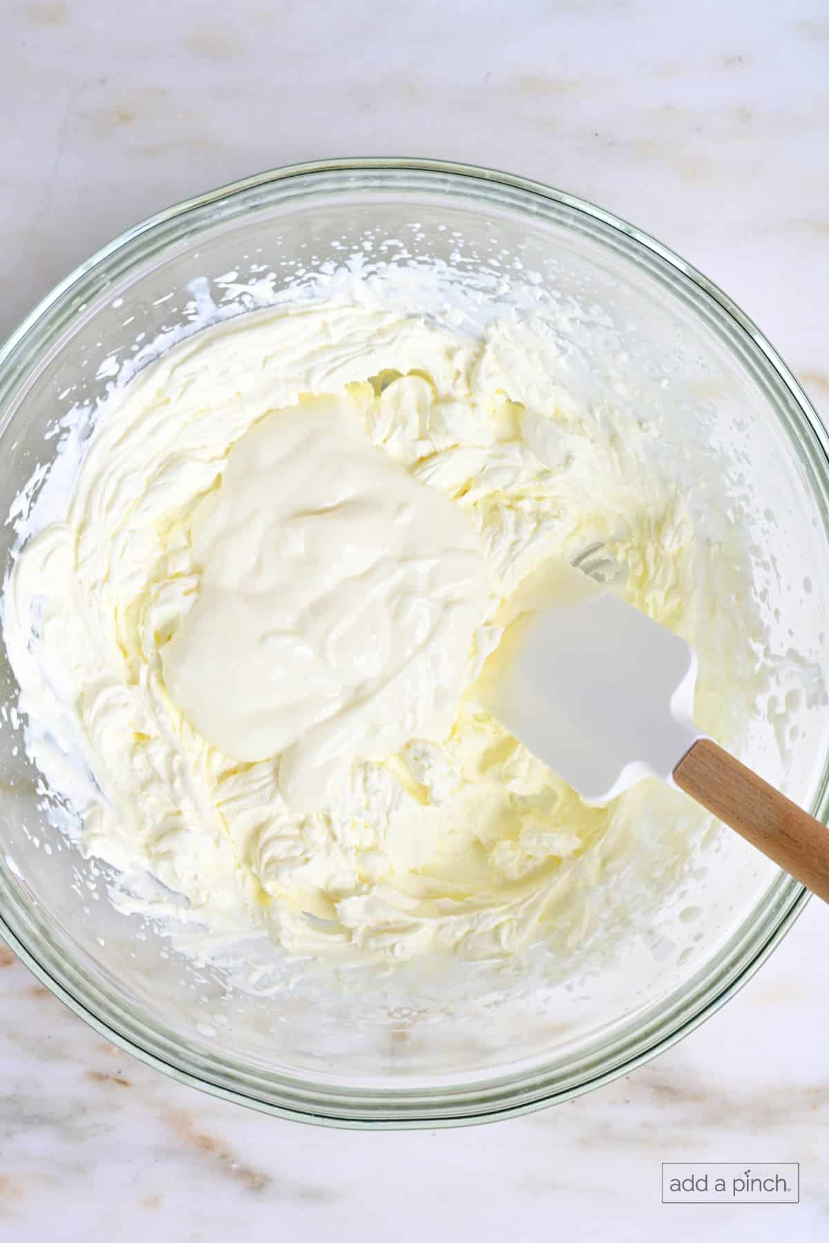 Whipped cream and yogurt in a glass mixing bowl with a rubber scraper.