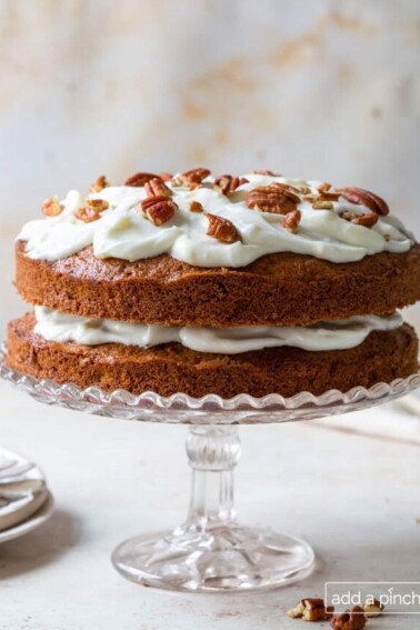 Carrot Cake recipe topped with cream cheese frosting, pecans on a clear glass cake stand.