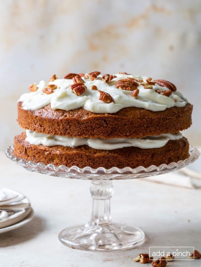 Carrot Cake recipe topped with cream cheese frosting, pecans on a clear glass cake stand.