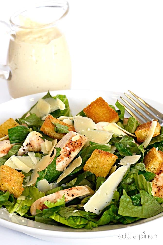 Chicken Caesar salad on a white bowl with a fork and dressing in the background. All on a white surface.