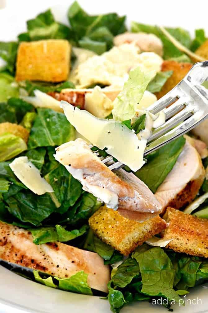 Bite of chicken caesar salad on a fork with full salad in a white bowl in the background.
