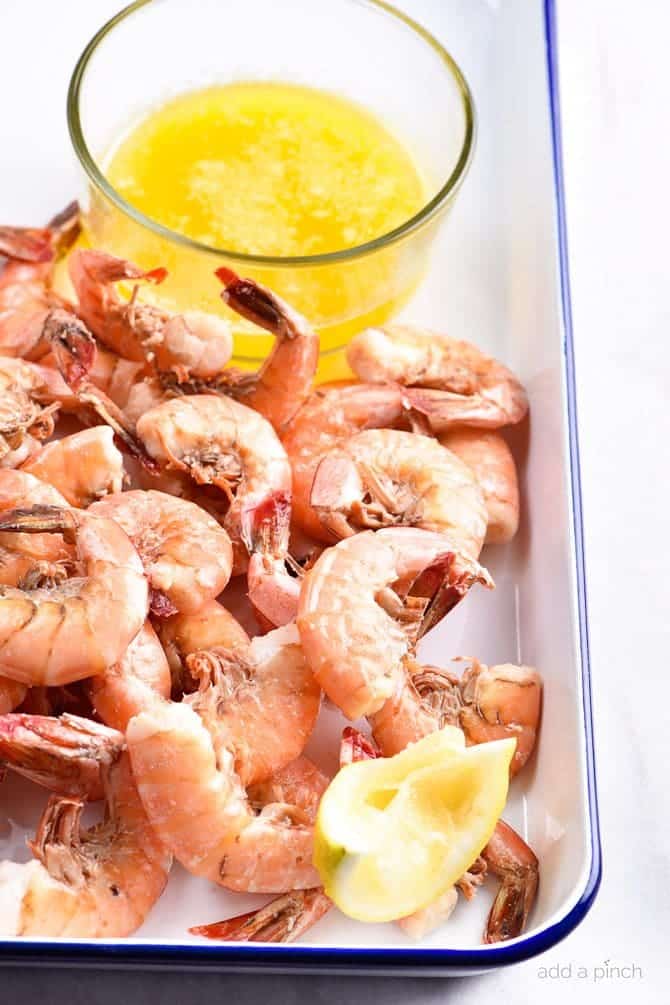 White platter with blue rim holds boiled shrimp with lemon wedge and glass container of garlic butter sauce.