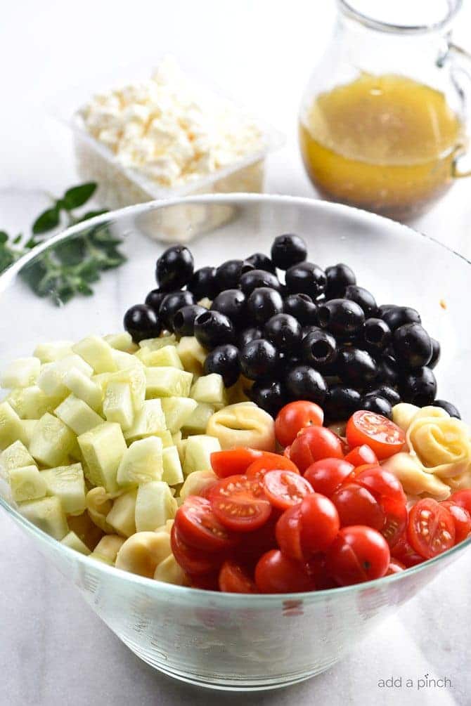 Glass bowl with chopped cucumber, sliced cherry tomatoes, and ripe black olives. Container of feta cheese, fresh oregano and glass dispenser with homemade Italian dressing on marble counter. // addapinch.com