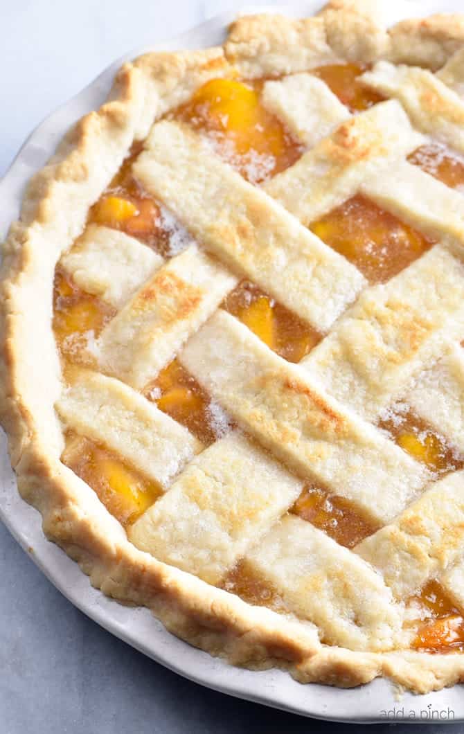 Photo of baked peach pie with a lattice crust in a white pie plate on a marble counter.