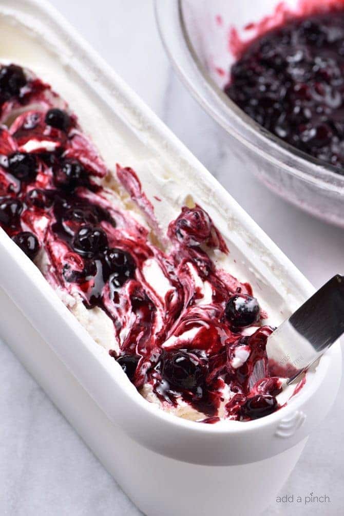 White tub of blueberry cheesecake ice cream with blueberry sauce swirl, beside bowl of blueberry sauce // addapinch.com