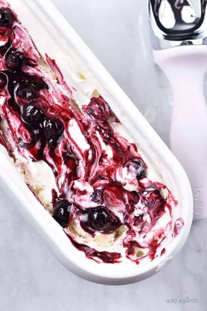 Tub of Blueberry Cheesecake Ice Cream with fresh blueberry sauce swirled on top, alongside a white and stainless ice cream scoop, all set on marble counter. // addapinch.com
