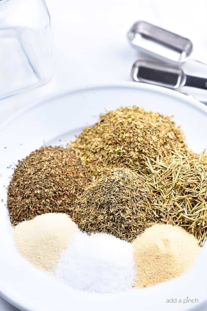 Dried herbs and spices are on a white plate, surrounded by measuring spoons.  