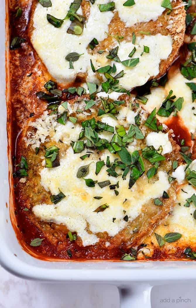 Closeup picture of eggplant layered with crispy baked eggplant, tomato sauce and cheese in a white baking dish. 