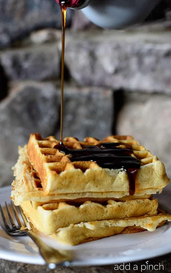 Our favorite buttermilk waffles stacked on a white plate with a fork and drizzled with a stream of syrup  // addapinch.com
