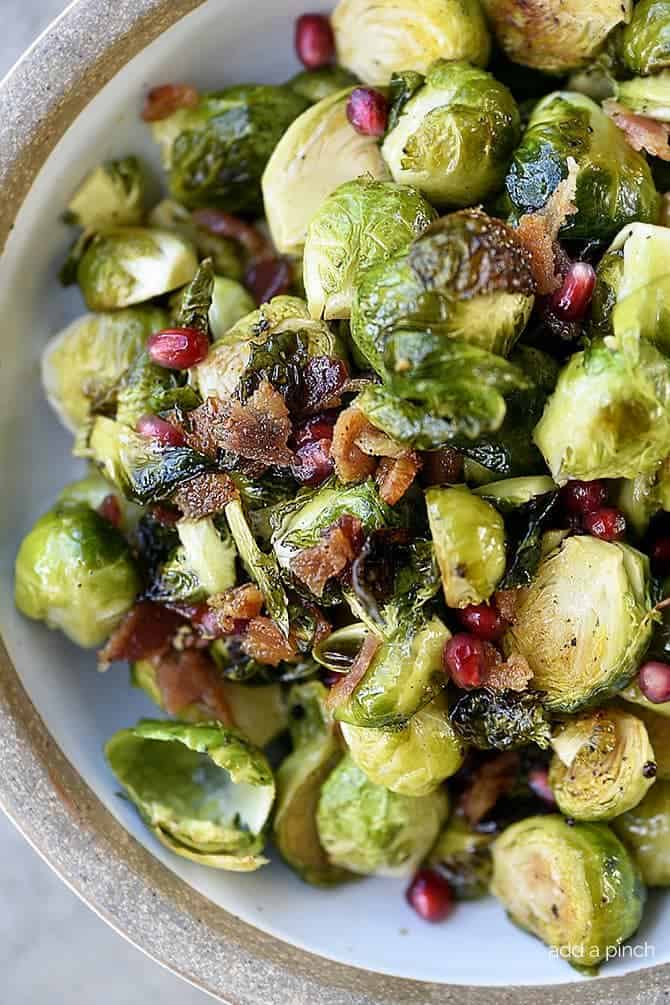 Roasted Brussels Sprouts are served in a white bowl mixed with bright pomegranate arils and crispy bacon pieces. // addapinch.com