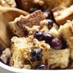 Baked Blueberry French Toast in a white baking dish is scooped with a wooden spoon, showing juicy blueberries and crisp streusel.