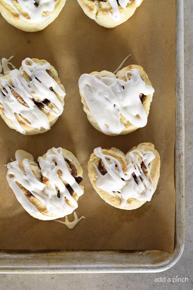 A parchment covered baking sheet holds several iced cinnamon roll biscuits made into heart shapes.