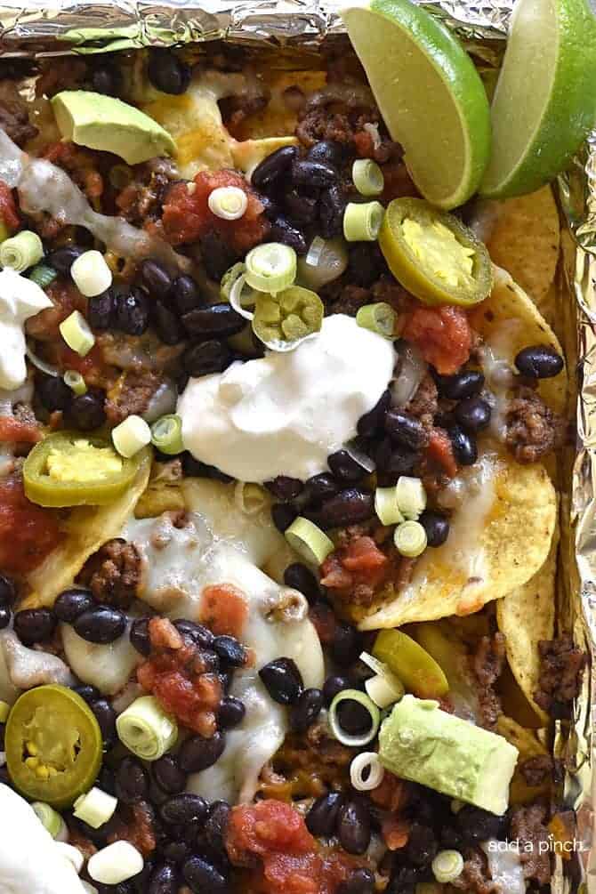 Side of sheet pan with nachos showing tortilla chips covered in melted cheese, beans, onions, jalapenos, salsa and slices of lime  // addapinch.com