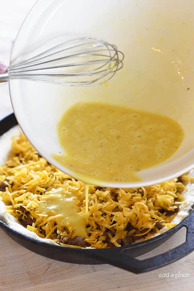 Egg mixture being poured from mixing bowl into iron skillet with biscuit crust, sausage, and cheddar. 