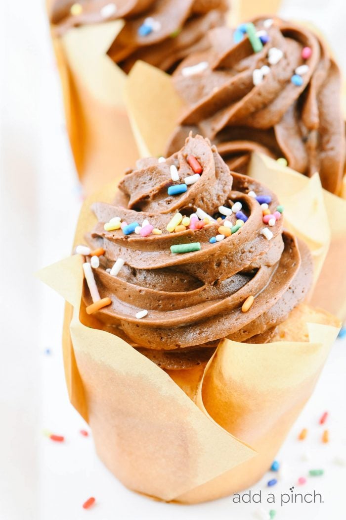 Closeup of cupcake topped with chocolate buttercream frosting and sprinkles in a parchment wrapper. 