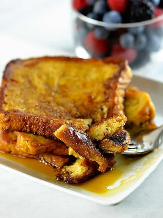 Photograph of French toast on a white plate with maple syrup and a bowl of fruit in the background.
