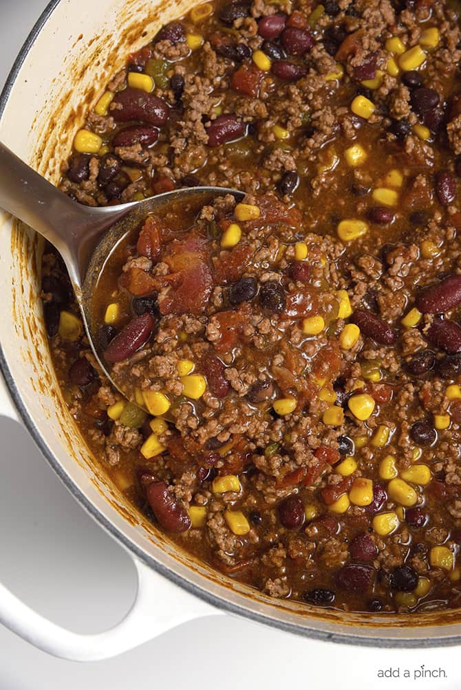 Ladle filled with meaty soup in a white Dutch oven with ground beef, corn, tomatoes and beans. 