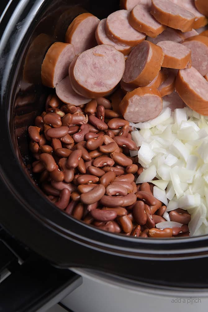 Slow cooker insert filled with red beans, chopped onion, sliced sausage  // addapinch.com