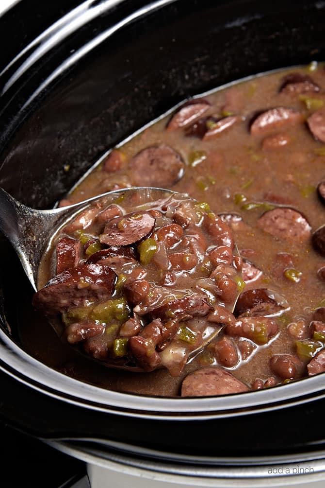 Slow Cooker with cooked red beans recipe with sausage, bell pepper and onions all spooned into a ladle // addapinch.com