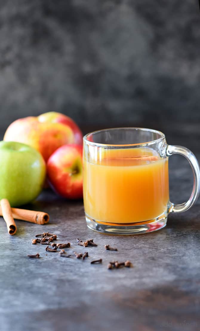 Cider in glass mug is surrounded by cinnamon sticks, cloves and apples. 