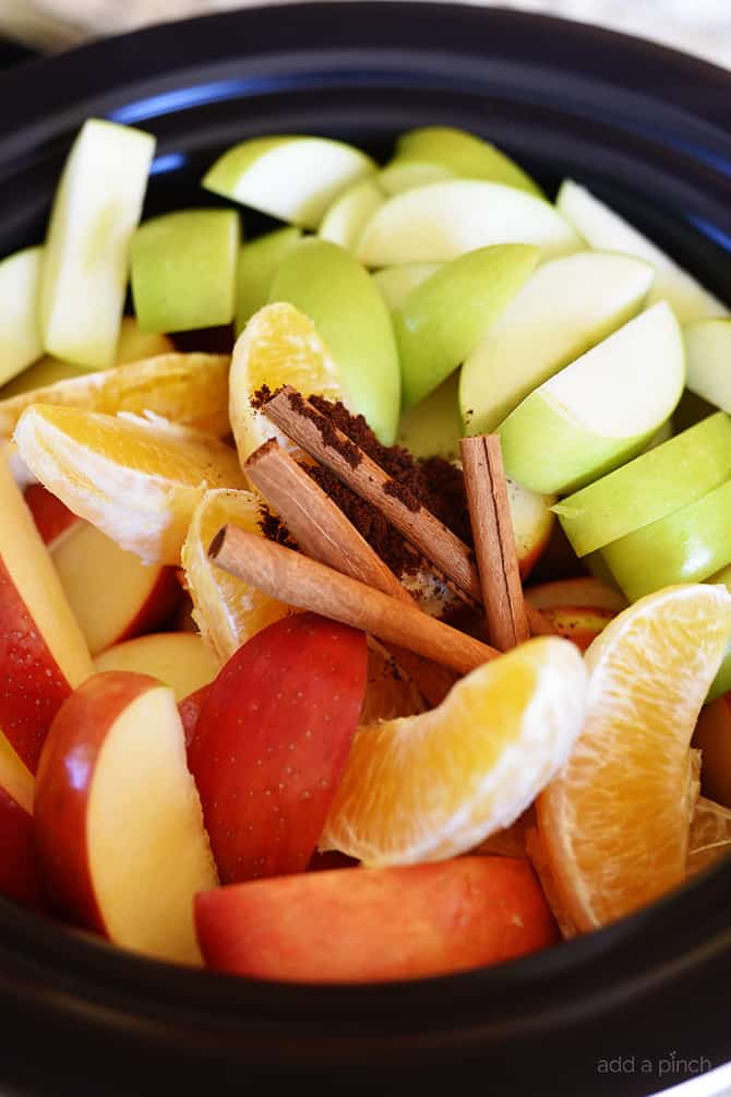Photograph of fresh red and green apple slices, orange slices, spices, and cinnamon sticks in a slow cooker insert. 