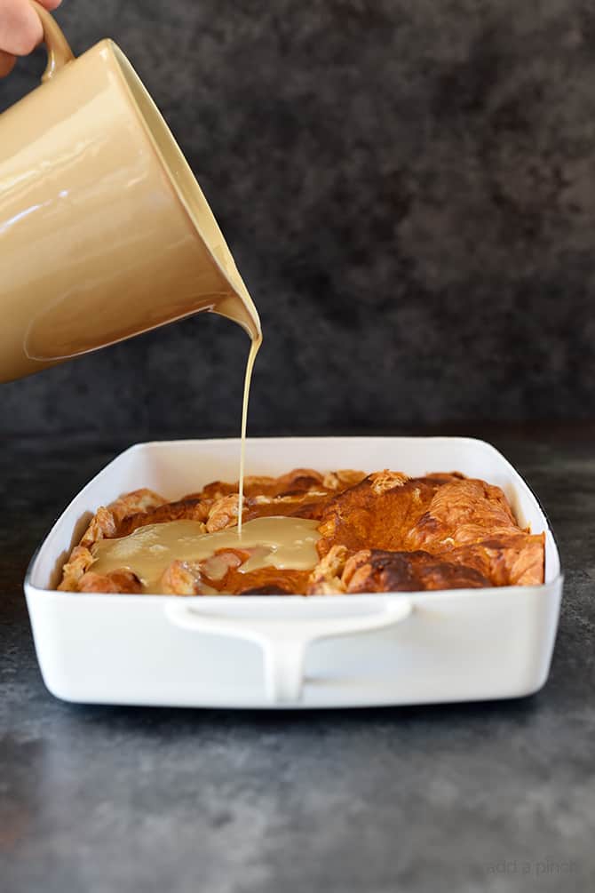 Maple cream sauce being drizzled from beige crockery pitcher onto pumpkin dessert in a white baking dish. 