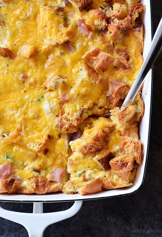 A scoop of golden brown, cheesy ham and cheese croissant casserole is being spooned out of a white baking dish.
