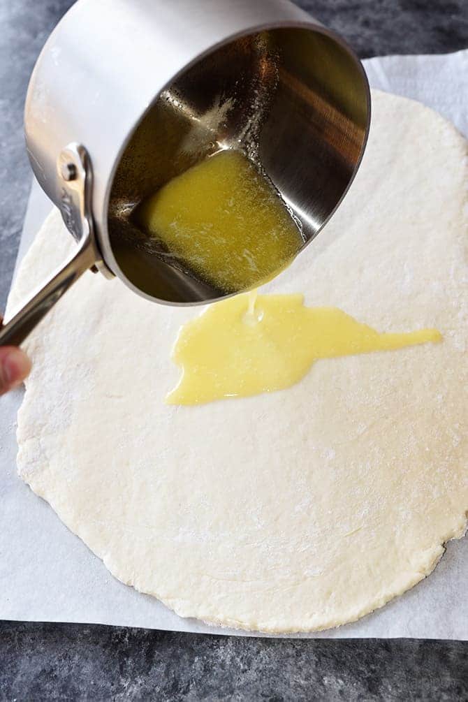 Stainless pot of melted butter being poured onto rolled biscuit dough.