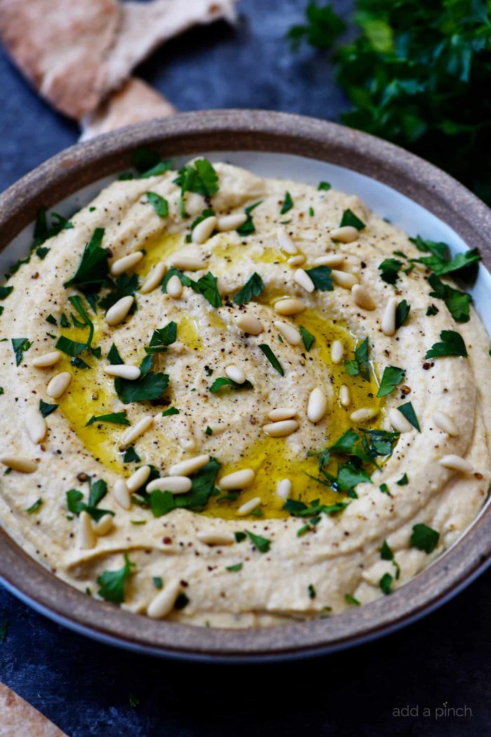 Bowl of hummus with pita bread in background, as well as parsley/ addapinch.com