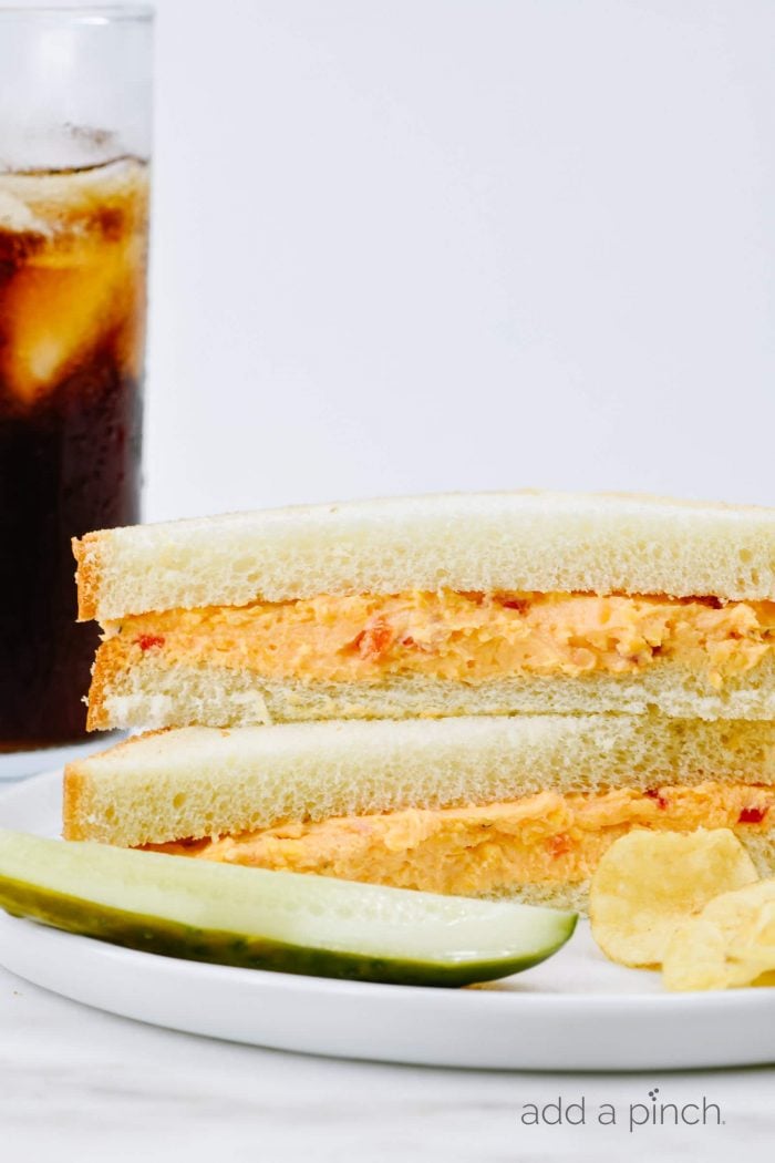 Pimento cheese sandwich on white bread is served on a plate with dill pickle spear and potato chips, along with a glass of iced sweet tea. 