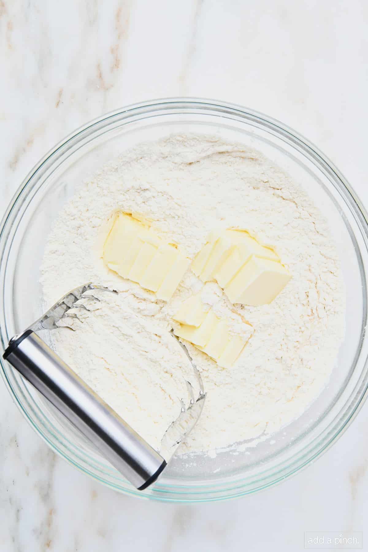 Butter being cut into flour mixture in a glass bowl with a pastry blender. 