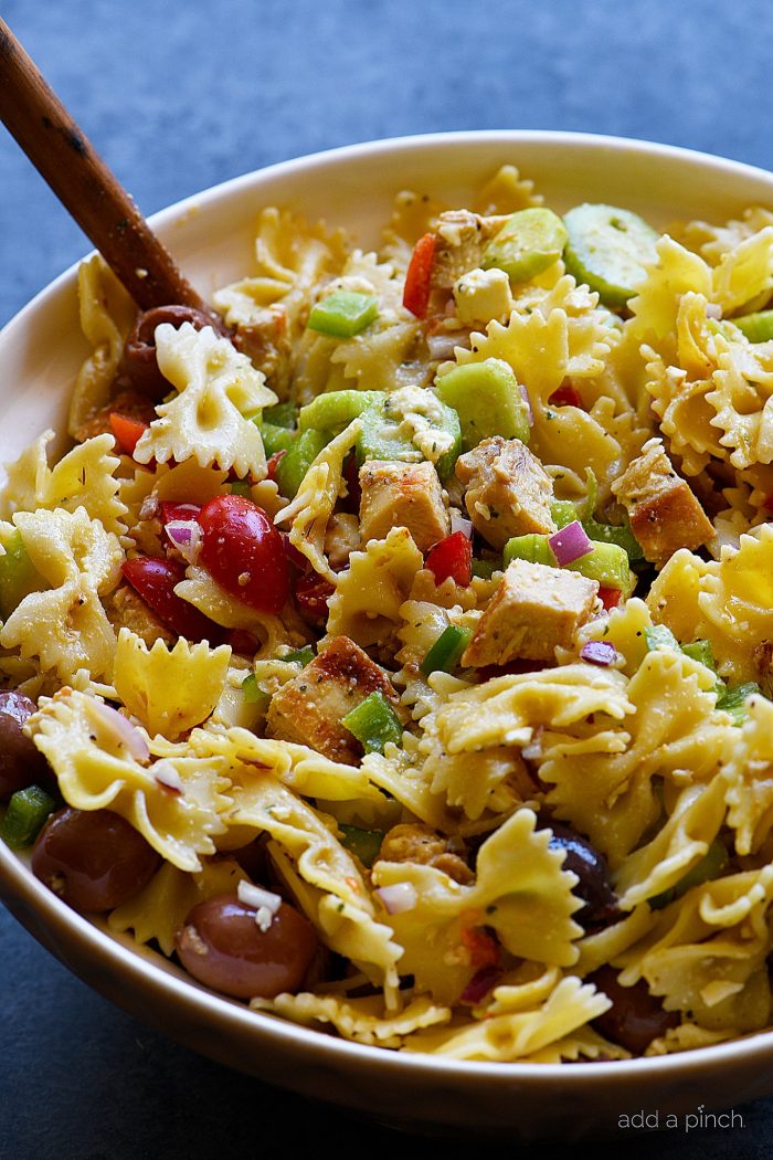 Wooden spoon stirs farfalle pasta and grilled chicken, an assortment of vegetables, olives and feta cheese 