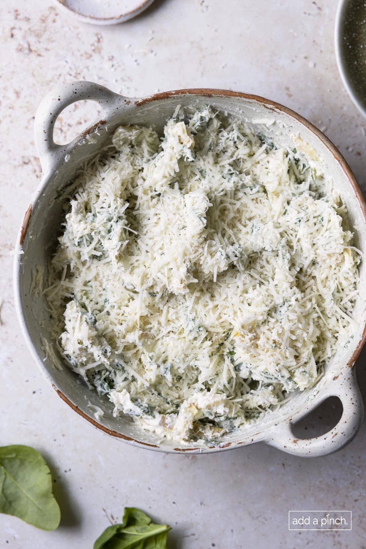 Dip  in round baking dish is covered in shredded parmesan and ready to be baked.