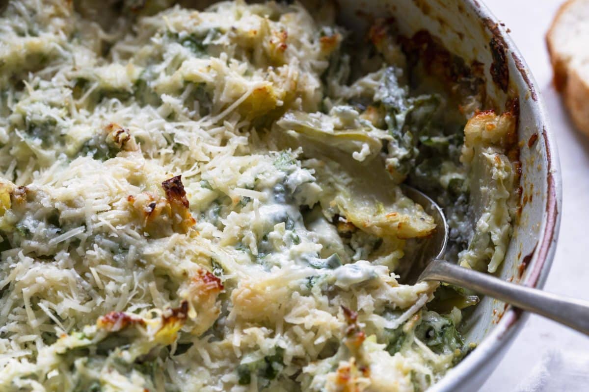 Photograph of baked spinach artichoke with a serving spoon in a white baking dish.