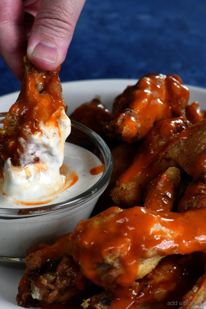 Chicken wings in buffalo sauce sit on piled on white plate with glass cup of homemade ranch dressing.