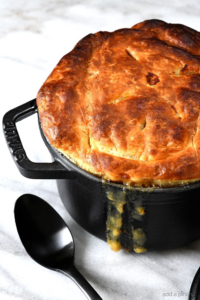 Black dutch oven is filled with chicken pot pie topped with a gold brown puff pastry top with thick juices running down the side. Sitting on marble countertop with black serving spoon. 
 // addapinch.com