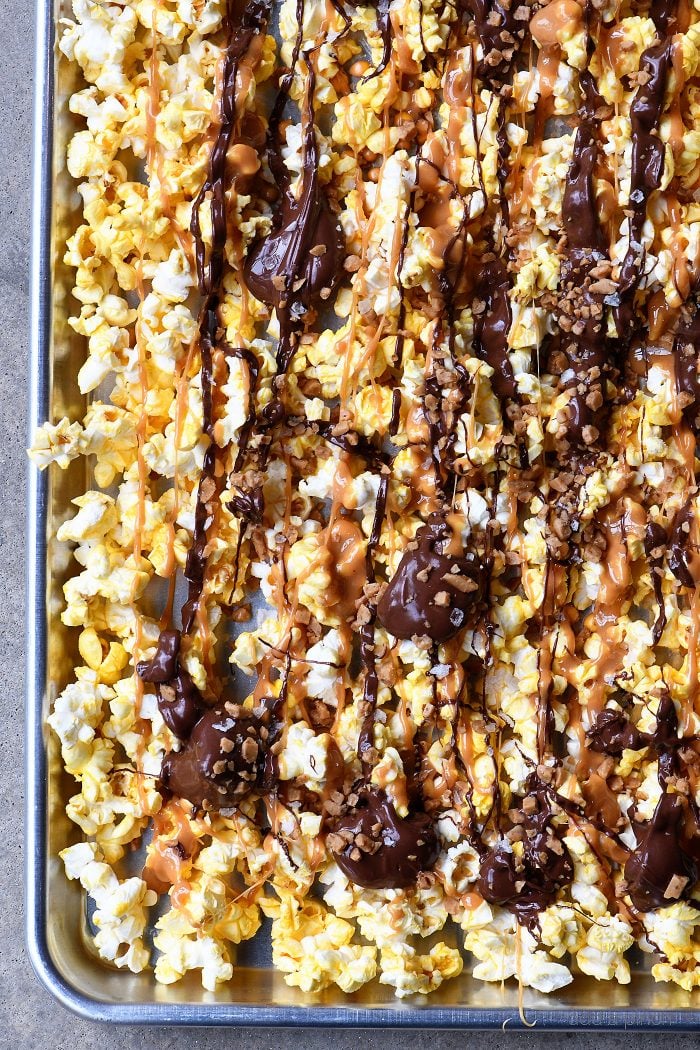 Caramel and chocolate are drizzed onto popcorn on a baking sheet and topped with toffee bits // addapinch.com