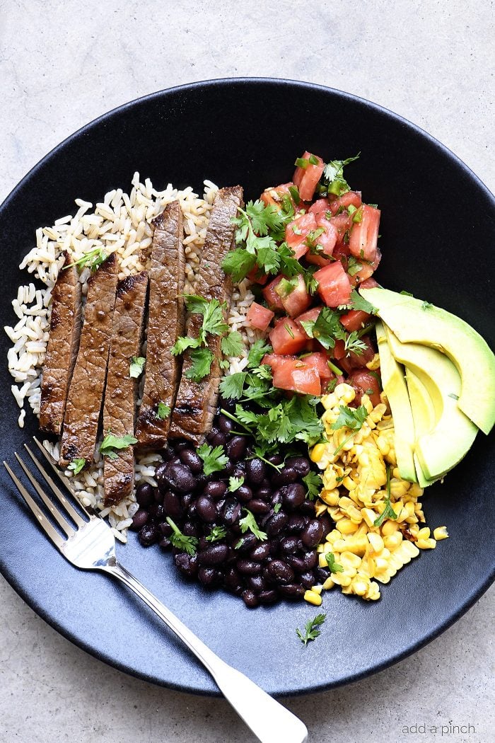 Dark bowl with rice, strips of steak, corn, black beans, avocado slices, pico de gallo, and cilantro