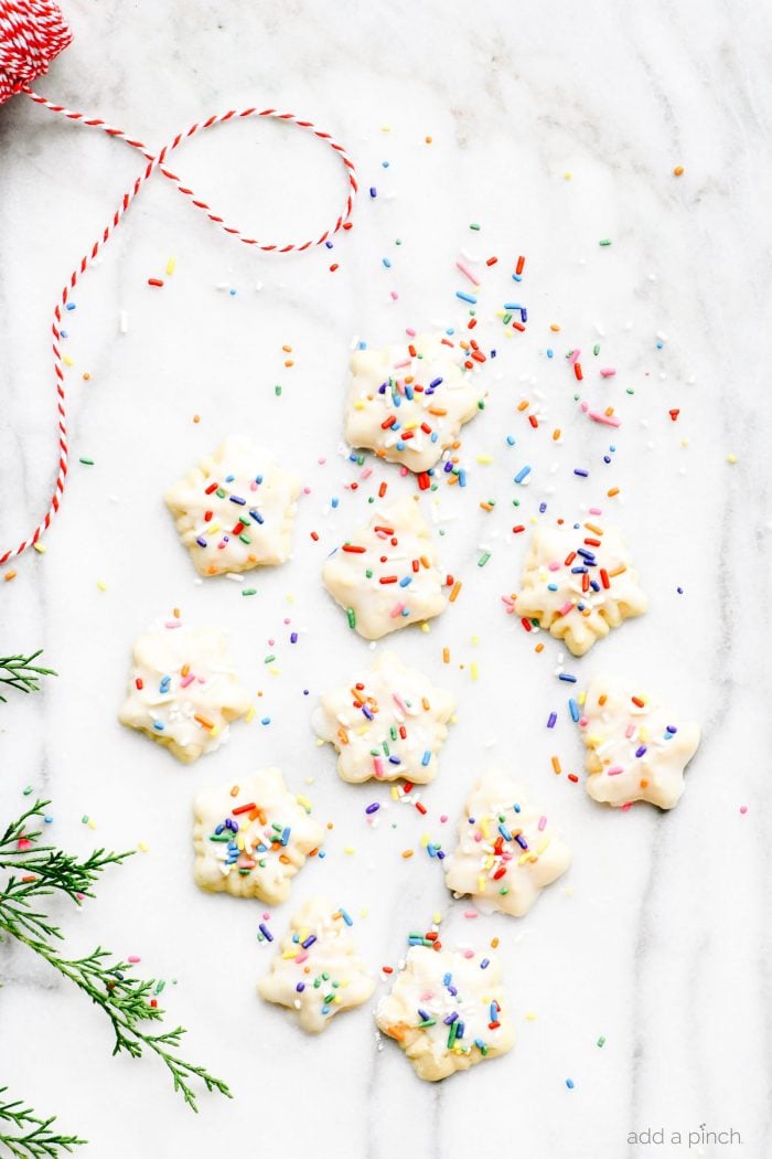 Spritz cookies glazed with multicolor sprinkles on a marble countertop surrounded by greenery and red and white twine