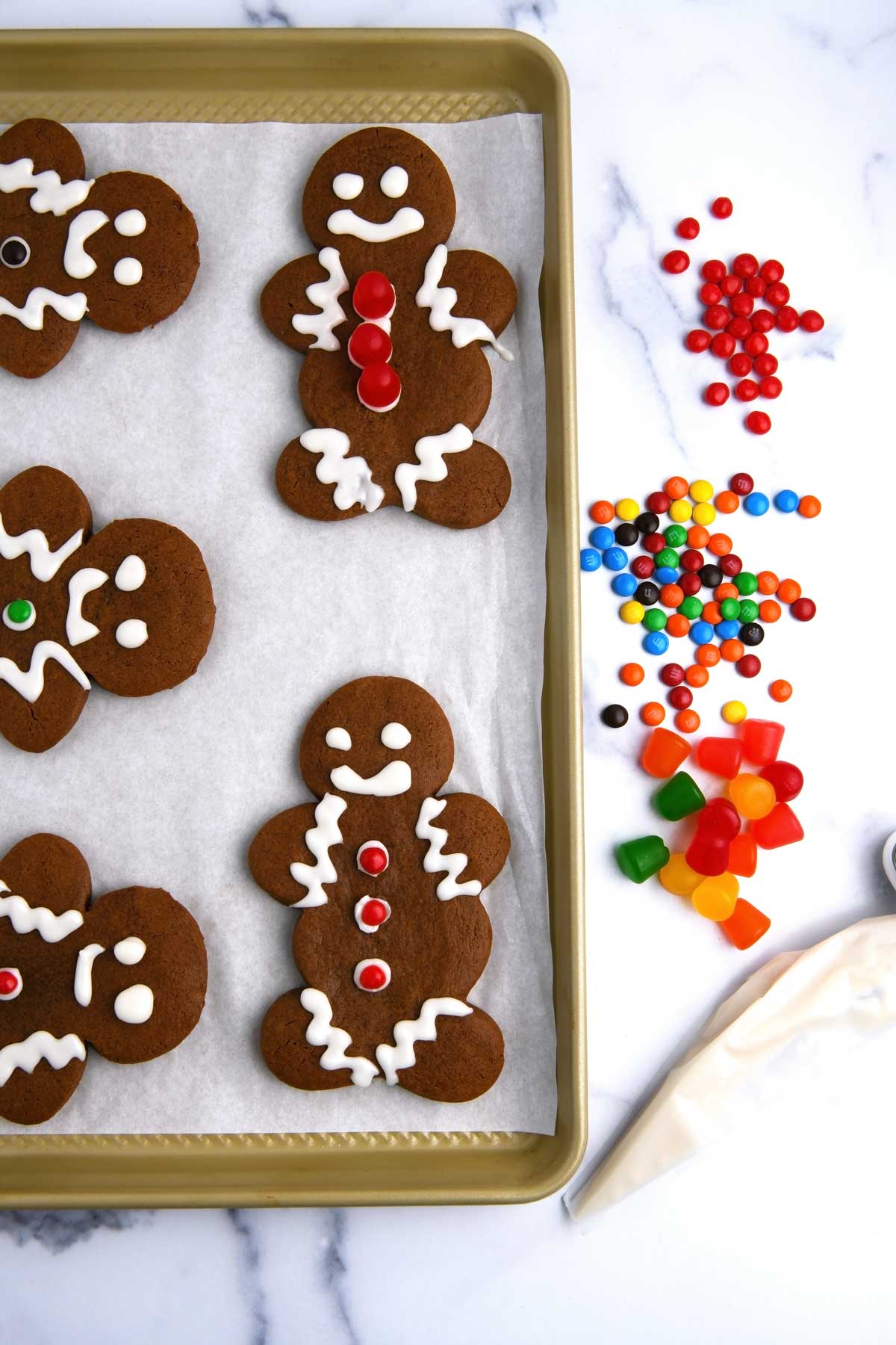 Photo of decorated gingerbread men cookies on a baking sheet with a frosting bag filled with cookie icing and assorted candy decorations on a marble countertop.