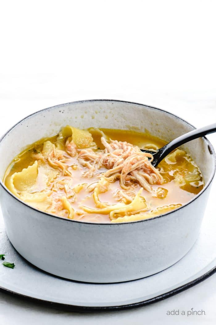 Photograph of a bowl of chicken noodle soup on a black rimmed plate on a white marble.