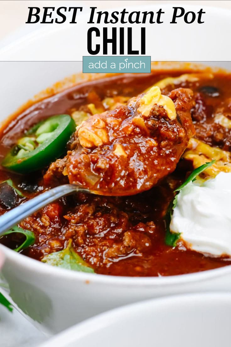 Bowl of Best Instant Pot Chili topped with cheese, sour cream, jalapeno slices with spoon - with text - addapinch.com