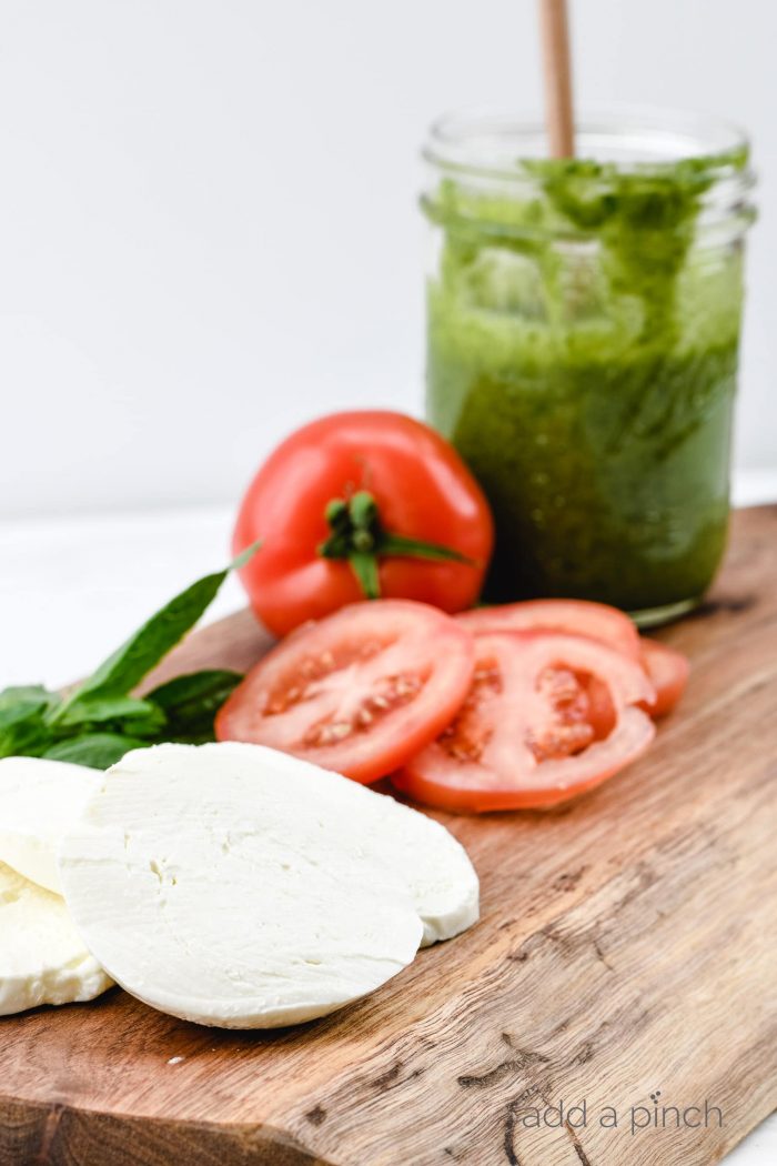 Sliced ripe tomato, jar of homemade pesto, slices of mozzarella and fresh basil leaves are set on wooden board // addapinch.com