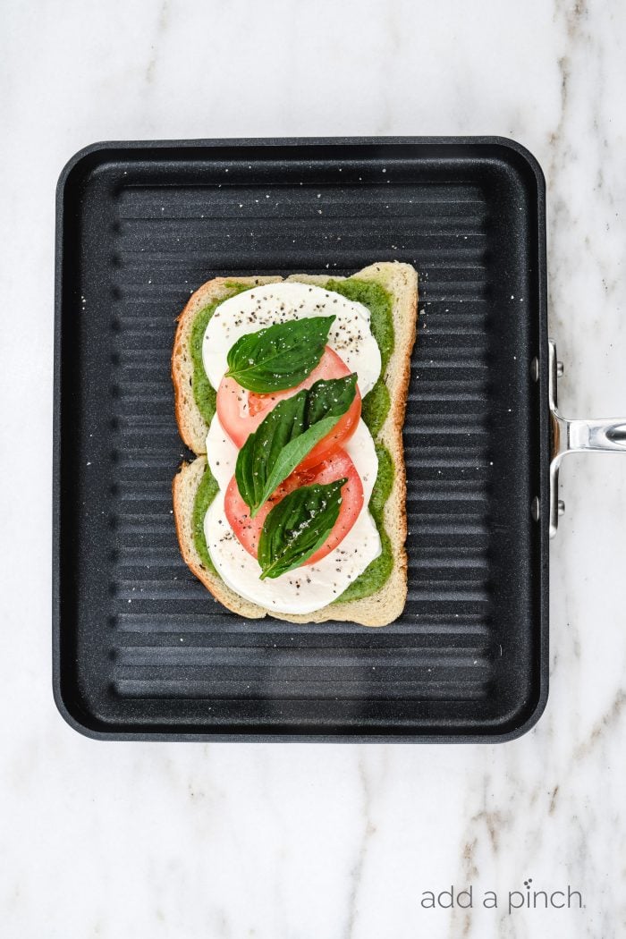 An assembled Caprese Grilled Cheese sandwich sits on grill pan showing layer of pesto, mozzarella, tomatoes and basil leaves // addapinch.com