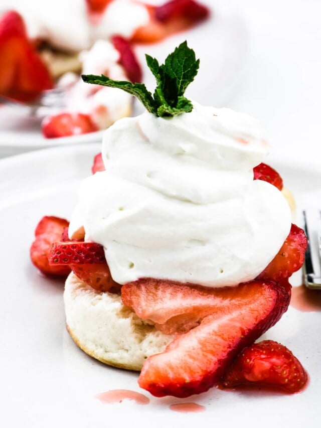 Strawberry shortcake topped with homemade whipped cream and mint sprig on a white plate.
